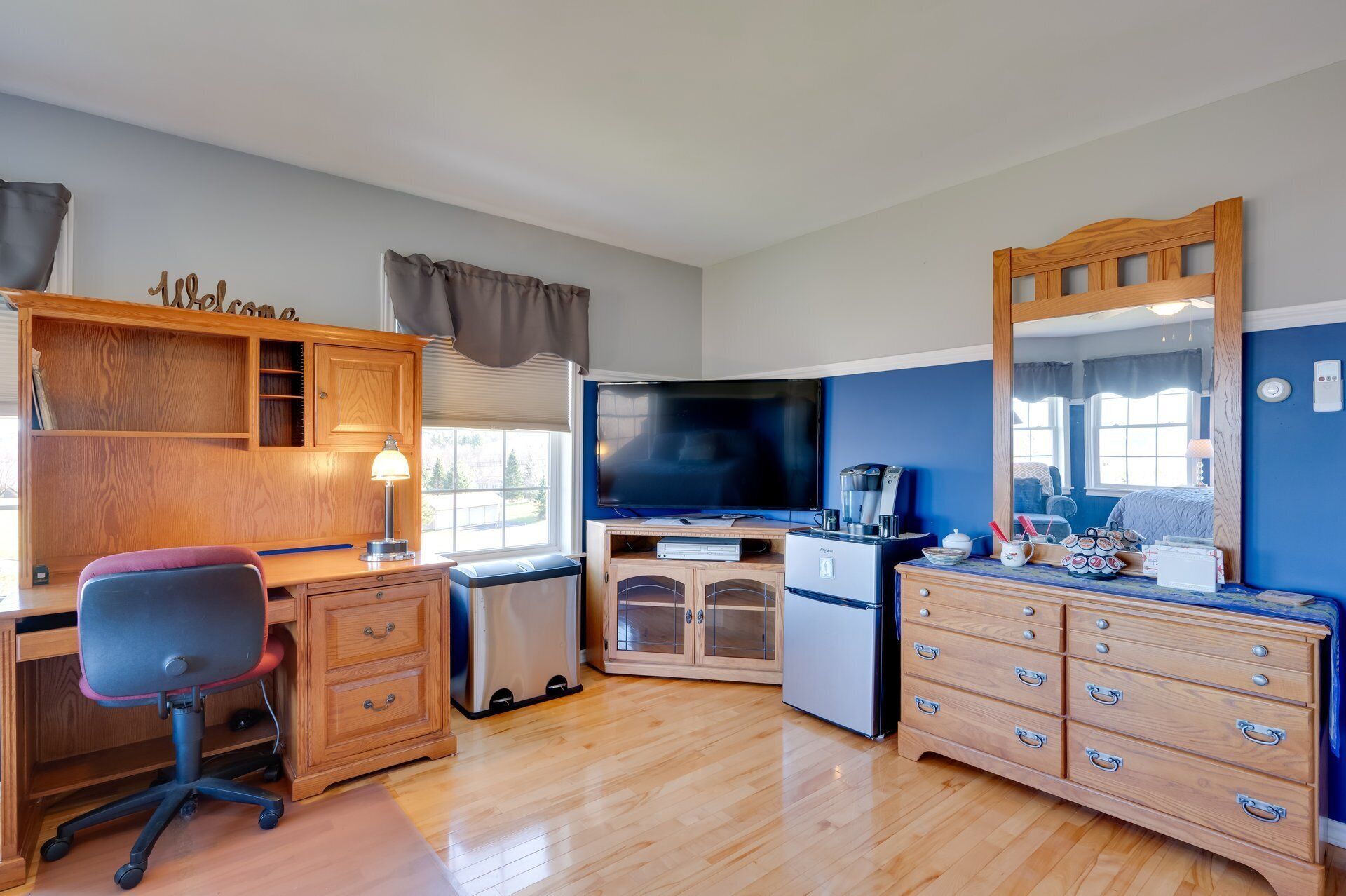 A compact room with a wooden desk, a chair, a television, a kitchenette, a dresser, and a mirror.