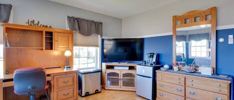 A compact room with a wooden desk, a chair, a television, a kitchenette, a dresser, and a mirror.