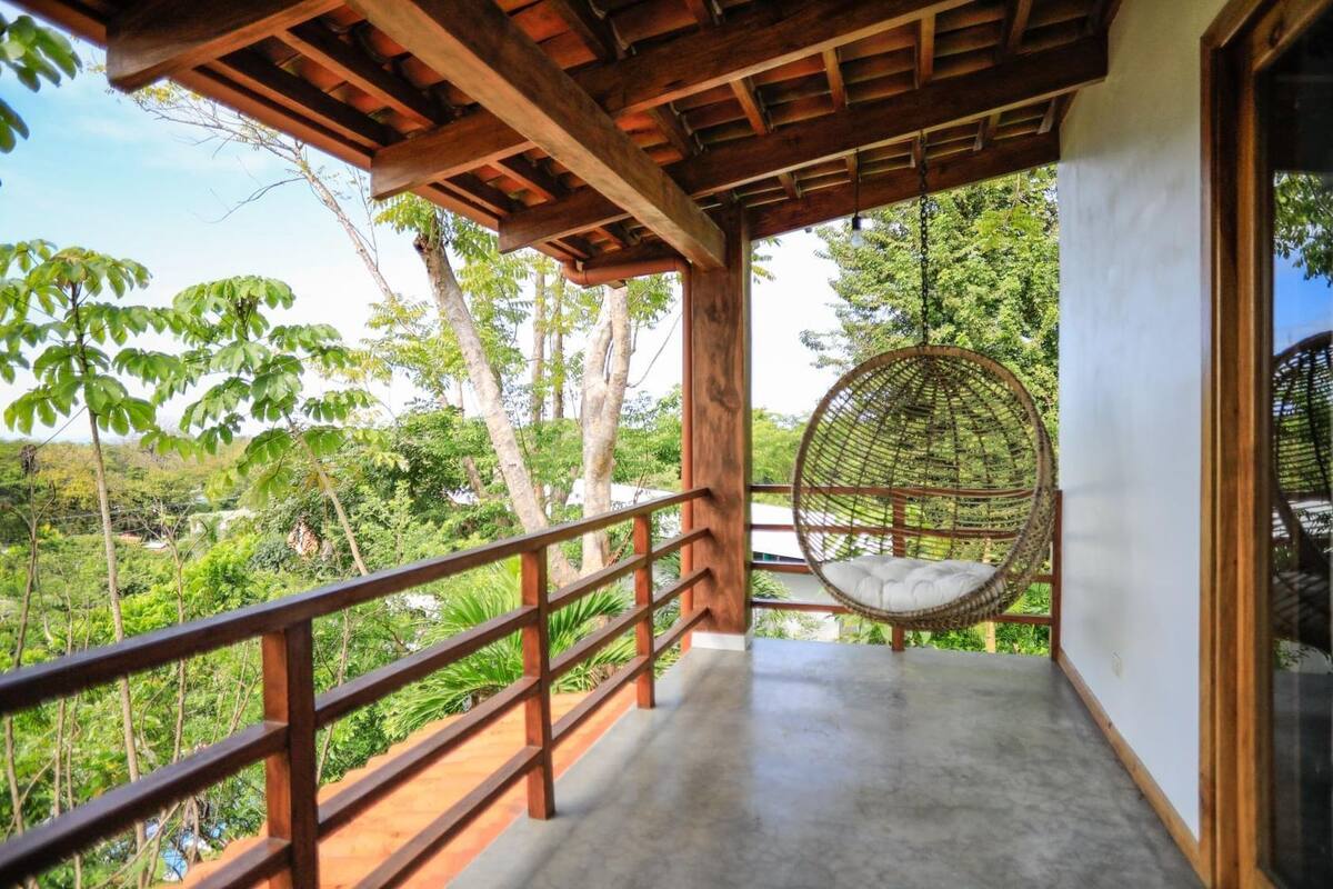 A balcony with a hanging chair, wooden railings, and a view of a lush green landscape.