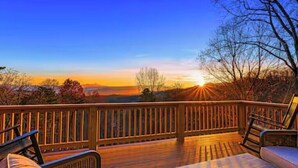 A wooden deck with a railing, two cushioned chairs, and a view of the sunset over a landscape. - Blue Ridge Cabin w/ Gorgeous Mountain Views! (Candler)