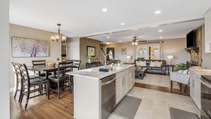 A modern kitchen with a central island, dining area with chairs, a sofa, and a TV mounted on the wall. - Blue Ridge Cabin w/ Gorgeous Mountain Views! (Candler)