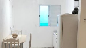 A small, compact room with a bed, a chair, a table, a window, and a refrigerator. - Pousada Caravela (Penha)