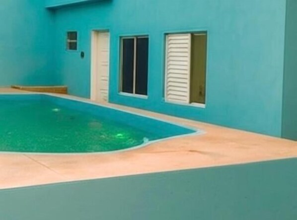 Une piscine dotée d’une plage de couleur claire, entourée d’un bâtiment aux teintes turquoises, avec des fenêtres et des portes à cadre blanc.