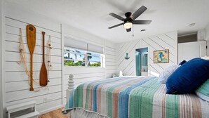 A bedroom with a bed, a ceiling fan, wooden paddles on the wall, and a window with a view. - Coral Way Getaway, Walk to Beach, Near Speedway, Sleeps 8 (Port Orange)