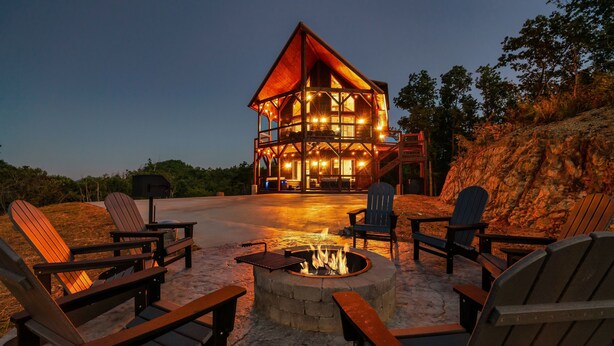 A lodge with a central fire pit surrounded by Adirondack chairs, a stone fireplace, and a wooden deck. - Smoky Mountain Cabin w/ Incredible View- Perfect for Families-Indoor Heated Pool (Sevierville)