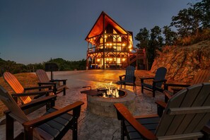 A lodge with a central fire pit surrounded by Adirondack chairs, a stone fireplace, and a wooden deck. - Smoky Mountain Cabin w/ Incredible View- Perfect for Families-Indoor Heated Pool (Sevierville)