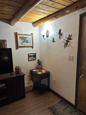Une pièce avec un plafond en bois, un téléviseur posé sur un meuble, une petite table recouverte d’une courtepointe, et des éléments de décoration murale comprenant des tableaux encadrés et des sculptures d’oiseaux.