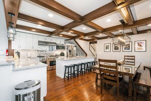 A spacious kitchen with wooden beams, a large island, bar stools, and a dining table with chairs. - 5BR Private Dock, Hot Tub, Firepit, Wisp (McHenry)