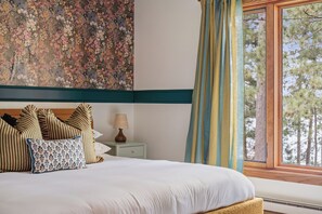 A neatly made bed with striped and patterned pillows, a bedside table with a lamp, and a window with a view of trees. - The Painted Turtle: Lakefront, Hot Tub, Sauna, Theater, Game Room, Playset (Nisswa)