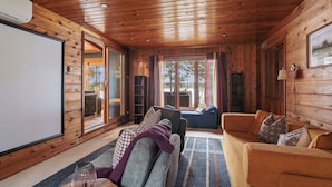Ein gemütliches Blockhauss-Interieur mit Holzbalkendecke, einem großen Fenster mit Vorhängen, einer Couch und einem Fernseher.