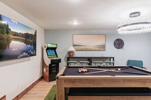 A pool table with balls, a large TV, a pool table game machine, and a chandelier. - The Painted Turtle: Lakefront, Hot Tub, Sauna, Theater, Game Room, Playset (Nisswa)