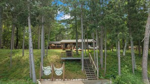A cabin surrounded by tall trees, with a wooden deck and a staircase leading to the entrance. - The Painted Turtle: Lakefront, Hot Tub, Sauna, Theater, Game Room, Playset (Nisswa)