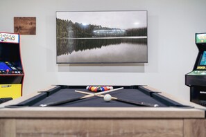 Et billardbord med et indrammet landskabsmaleri over, arkademaskiner til venstre og en poolkø i midten.