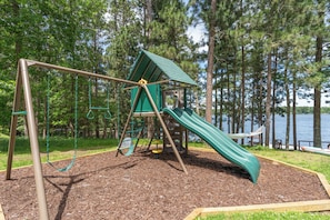 A playground with a slide and swings, surrounded by trees and a body of water. - The Painted Turtle: Lakefront, Hot Tub, Sauna, Theater, Game Room, Playset (Nisswa)