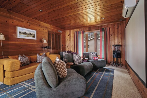 Et hyggeligt interiør i en hytte med træloft, et stort vindue med gardiner, en sofasæt og et lille bord med en lampe.