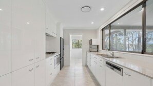 A modern kitchen with white cabinetry, stainless steel appliances, and a large window providing a view of trees. - Moonee Stayz – House - Where Moonee estuary meets the ocean (Moonee Beach)