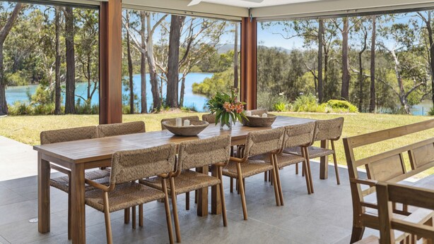 An outdoor dining area with a wooden table and chairs, a ceiling fan, and a view of a lake and trees. - Moonee Stayz – House - Where Moonee estuary meets the ocean (Moonee Beach)