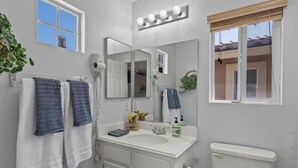 A bathroom with a white sink, a mirror with lights, a towel rack with towels, and a window with blinds.
