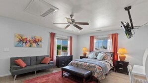 A bedroom with a bed, a sofa, a desk, a ceiling fan, and a TV mounted on the wall.