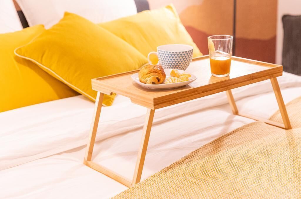 Een houten dienblad met een croissant, een kopje en een glas sinaasappelsap op een bed met gele kussens.