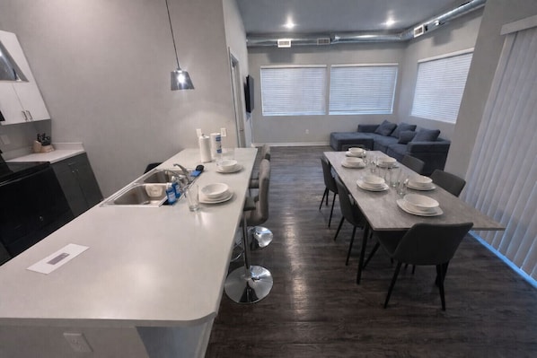 A modern kitchen with a central island, dining area with a long table, and a gray sofa in a compact living space.