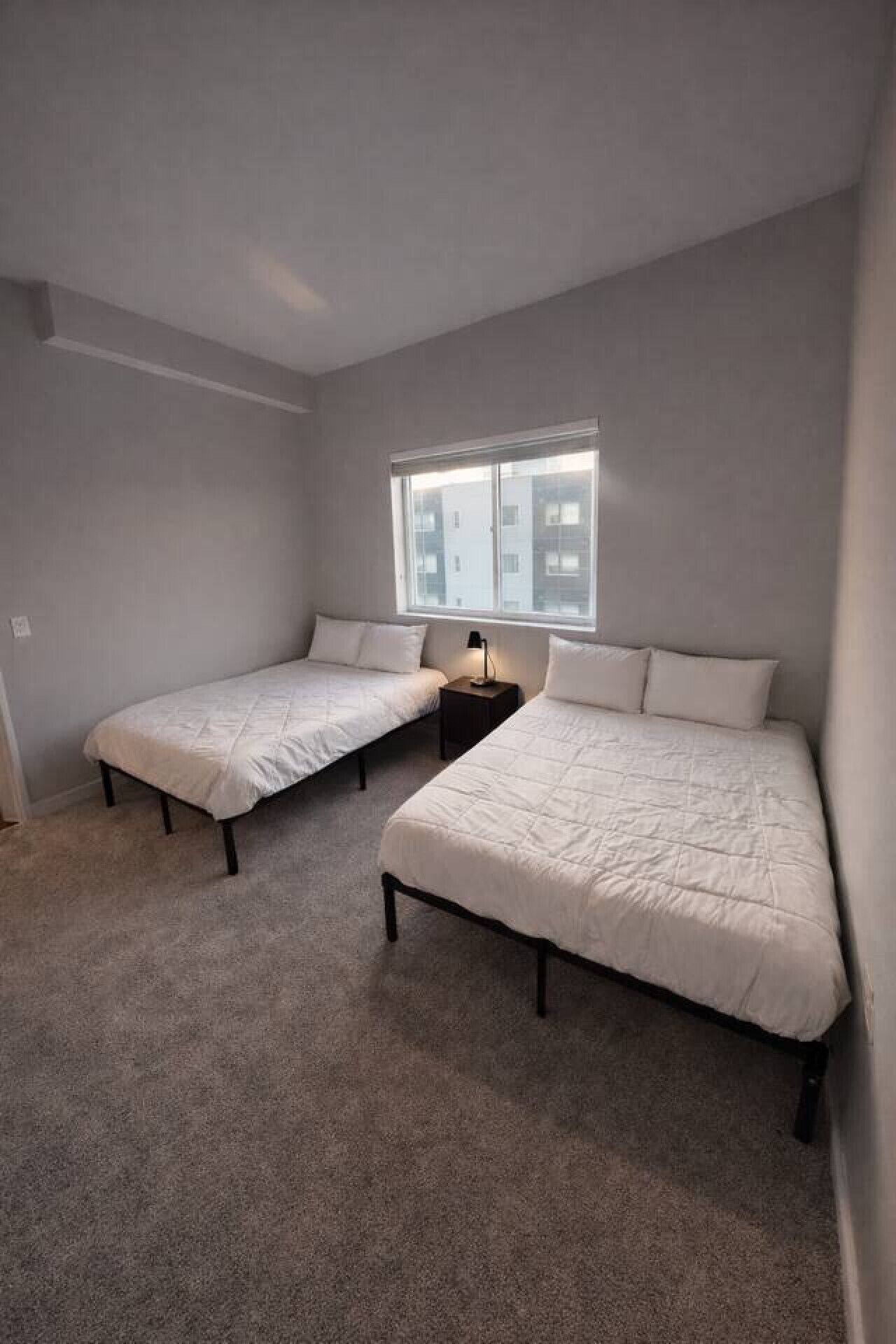 A room with two single beds, a small table with a lamp, and a window with a view of buildings.