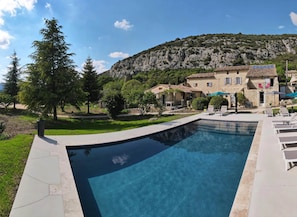 A swimming pool with a patio area, a stone building with a tiled roof, and a mountainous backdrop. - le Pitchounet, gite for 2 _ Lacets Ventoux (Beaumont-du-Ventoux)