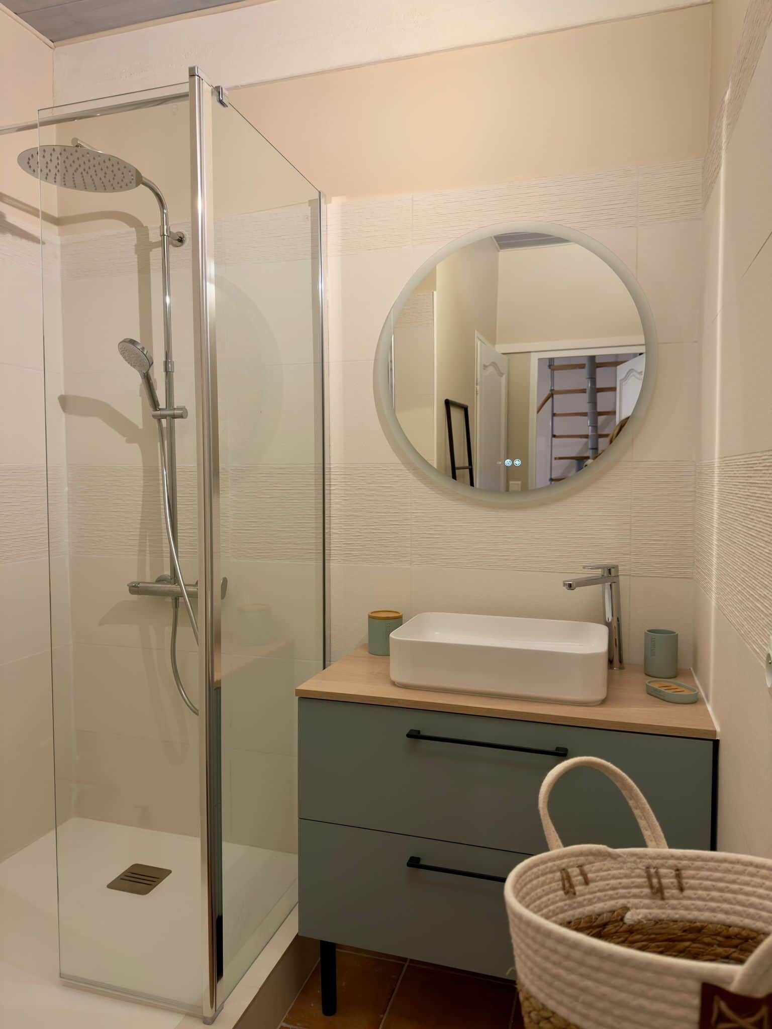 Un baño con cabina de ducha de vidrio, un espejo redondo, un lavabo con encimera de madera y una cesta.