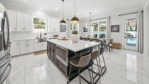 Una cocina moderna con armarios blancos, una isla grande con taburetes de bar, iluminación colgante y vistas al exterior.