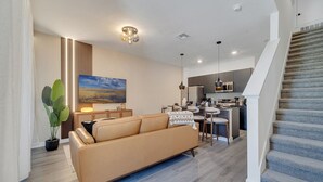 A modern living room with a sofa, a dining area, and a kitchen. - 3BR Home Near Disney w/ Office & Fun Corner (Kissimmee)
