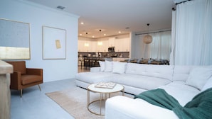 A modern living room with a sectional sofa, a round coffee table, and a brown armchair. There is a kitchen in the background with white cabinets and a dining area. - 6BR Family at Champions Gate Pool Game Room (Kissimmee)