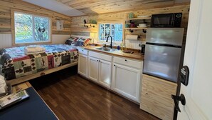 Een knus, huiselijk ingerichte kamer met een bed, kitchenette en een raam met uitzicht op bomen.