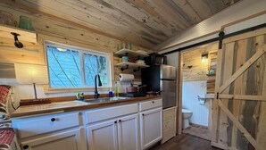 Een kleine keuken met witte kastjes, een houten aanrecht, een spoelbak en een fornuis. Er is een raam met uitzicht op bomen, en een schuifdeur die leidt naar een badkamer.