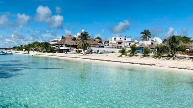 Una spiaggia con acque cristalline turchesi, una costa di sabbia bianca e edifici con tetti di paglia.