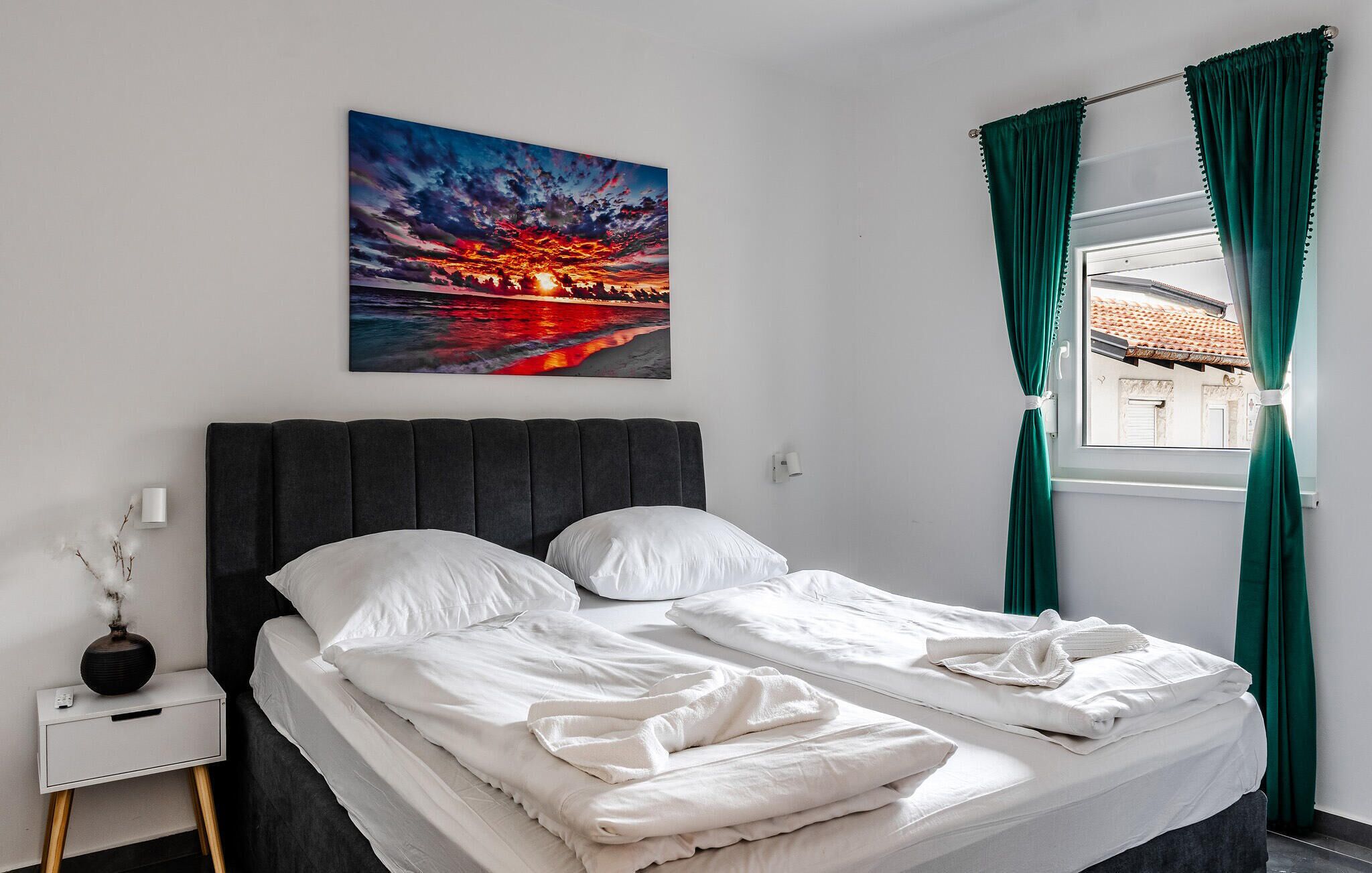 A neatly made bed with white linens, a dark headboard, and a painting of a sunset over water on the wall.