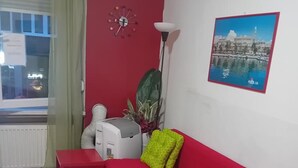En röd soffa med en grön kudde, en väggklocka, ett element, ett fönster med gardiner och en inramad tavla på väggen.