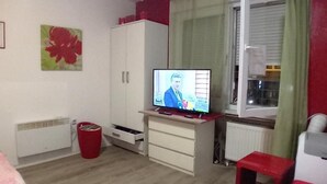 Ett rum med en tv, ett element, ett fönster med gardiner, en röd pall och en röd papperskorg.