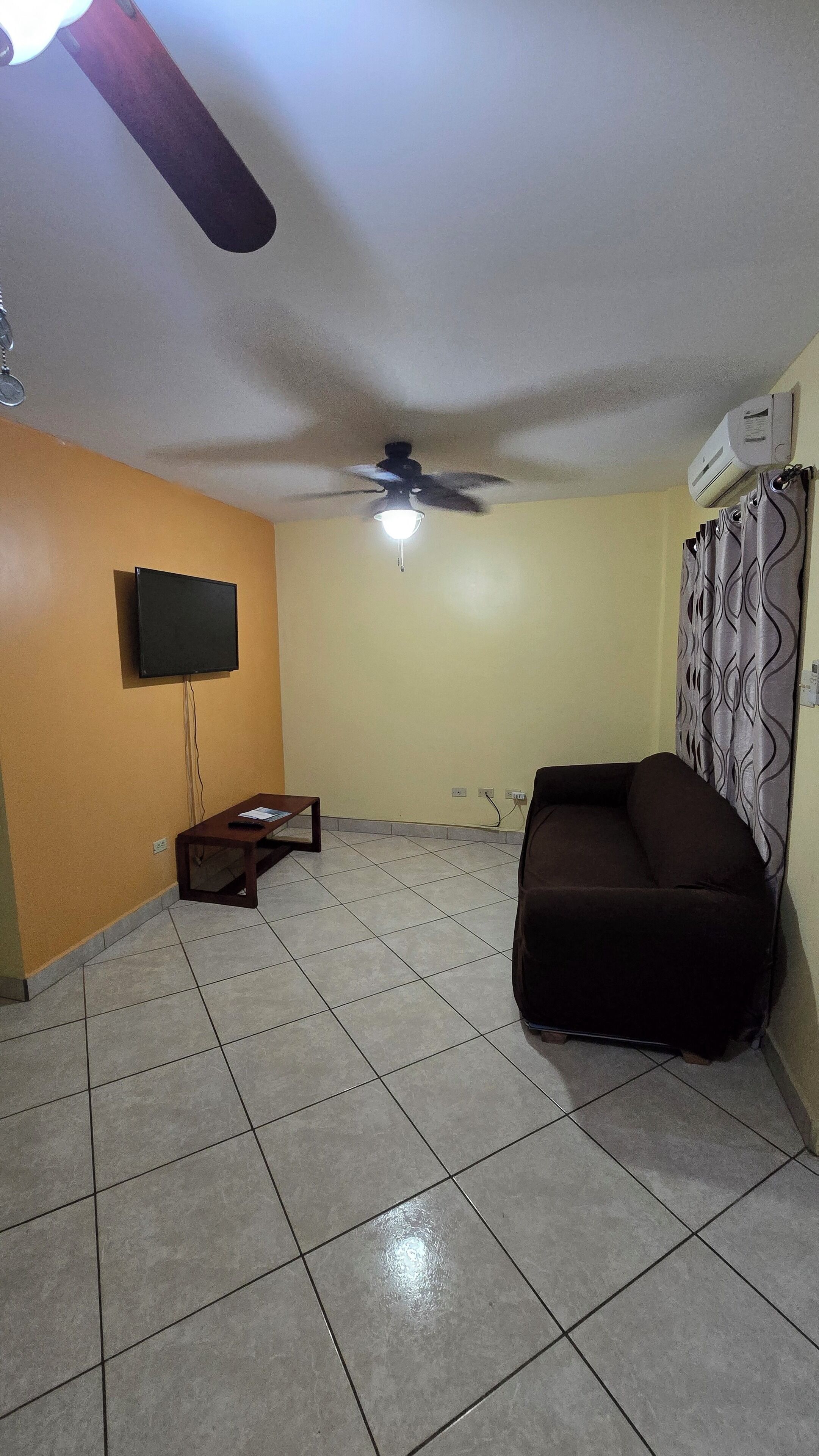 Una sala de estar con ventilador de techo, televisor de pantalla plana montado en la pared, sofá y una mesita con control remoto.