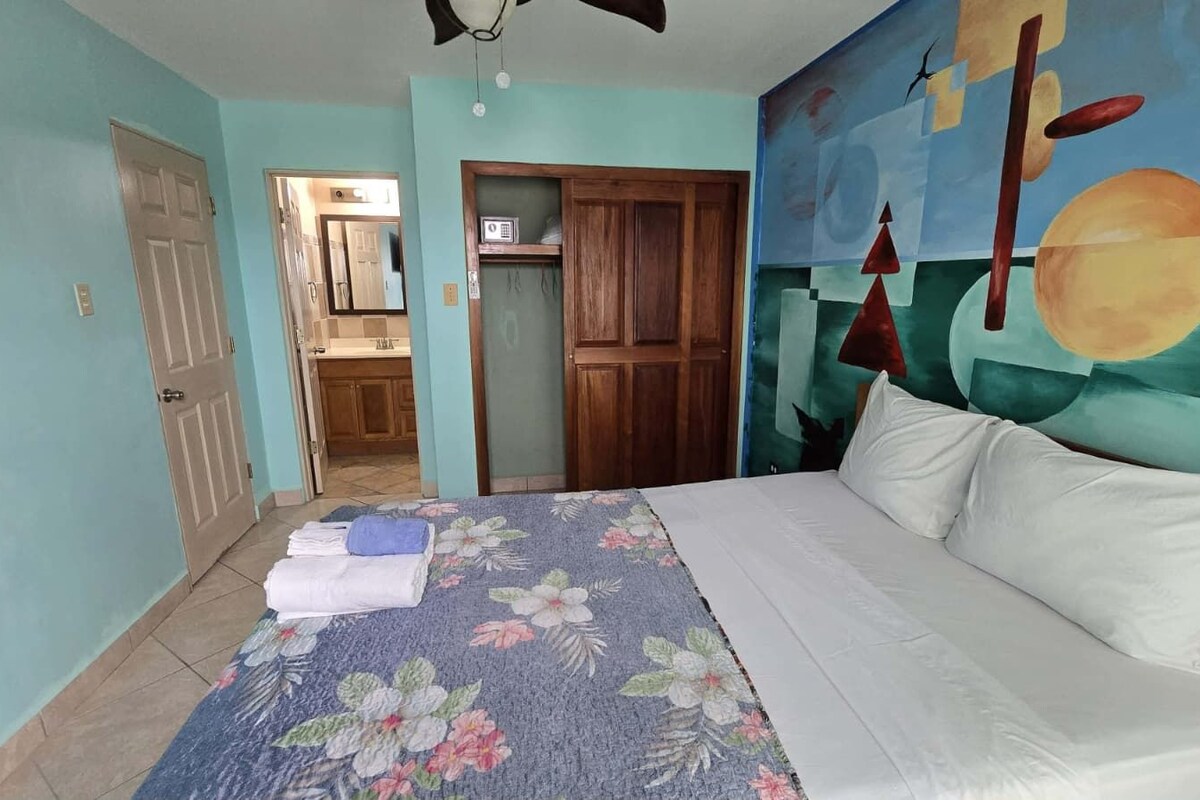 A bedroom with a bed, a floral bedspread, a wooden wardrobe, and a wall painting.