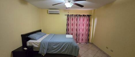 A bedroom with a bed, a ceiling fan, an air conditioner, a nightstand, and a curtain.