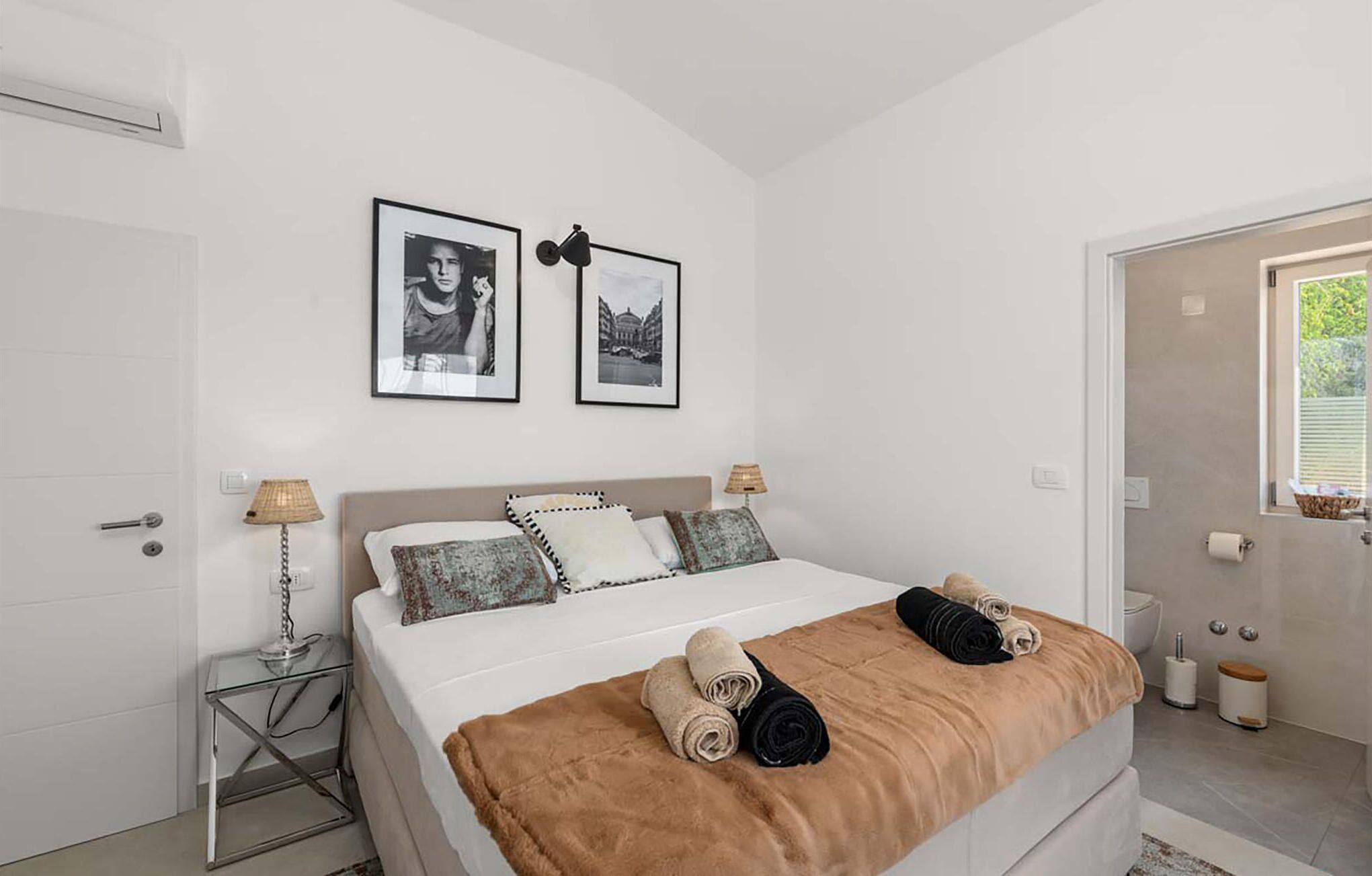A bedroom with a bed, two framed pictures, a bedside table with lamps, and a door with a window.