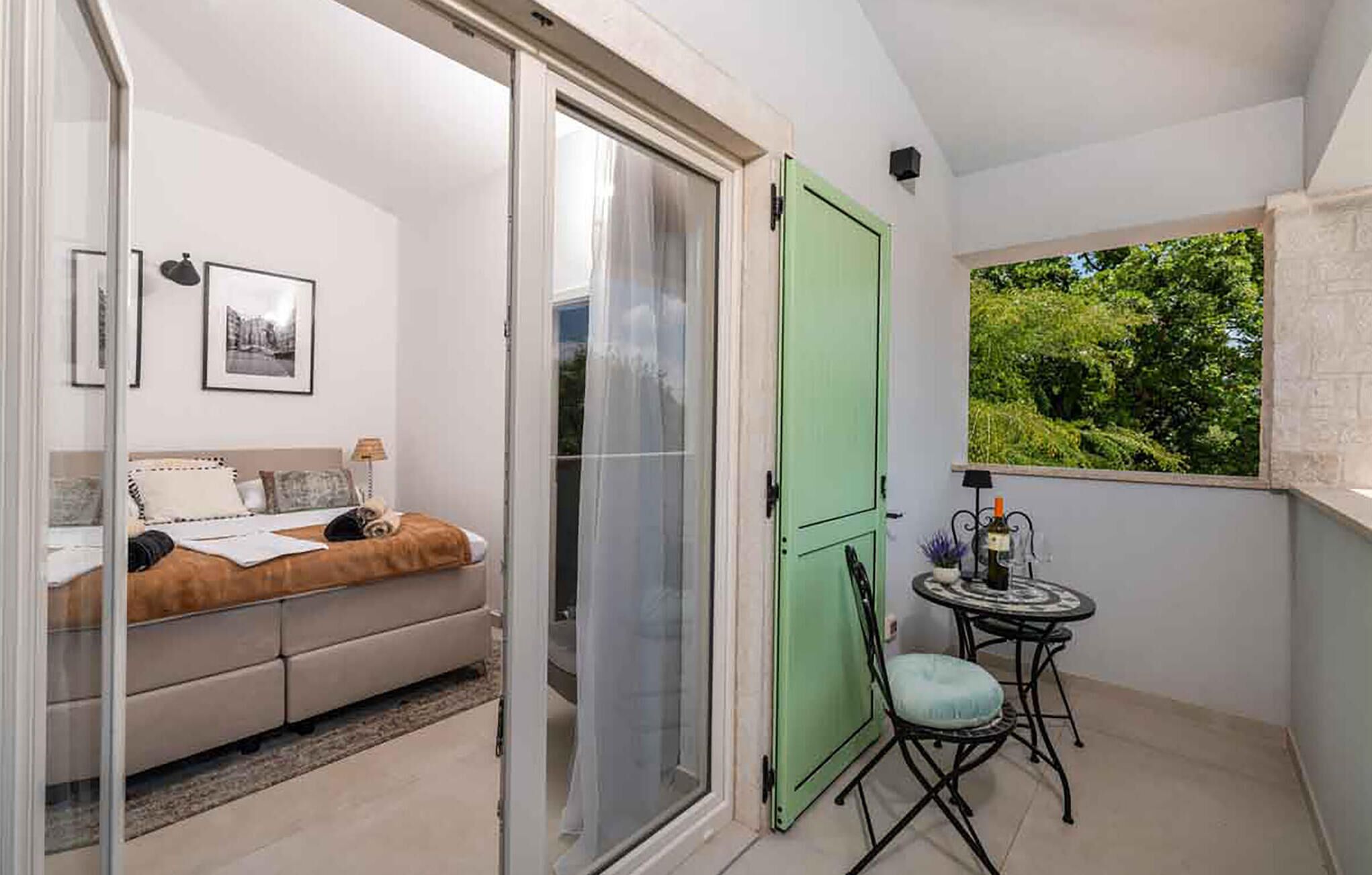 A balcony with a green door, a small table with a chair, a bed with a view, and a framed picture on the wall.