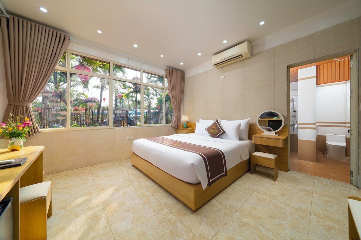 Habitación de hotel con una cama grande, un escritorio con silla, una mesita, un espejo y una ventana con cortinas.
