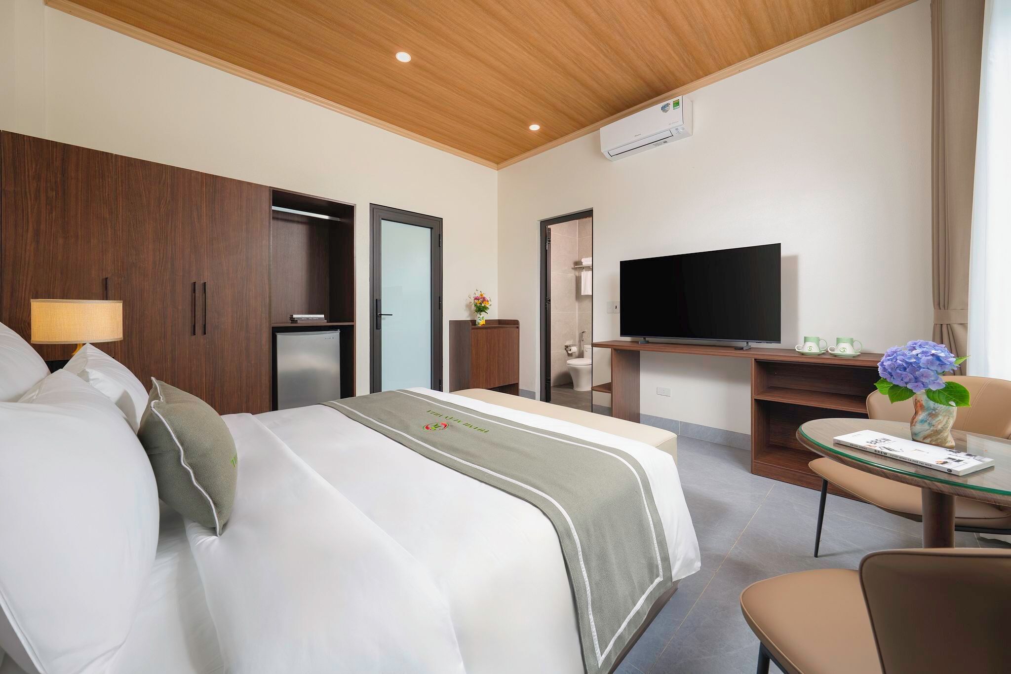 A hotel room with a bed, a desk with a flat-screen TV, a small table with a vase, and a chair.