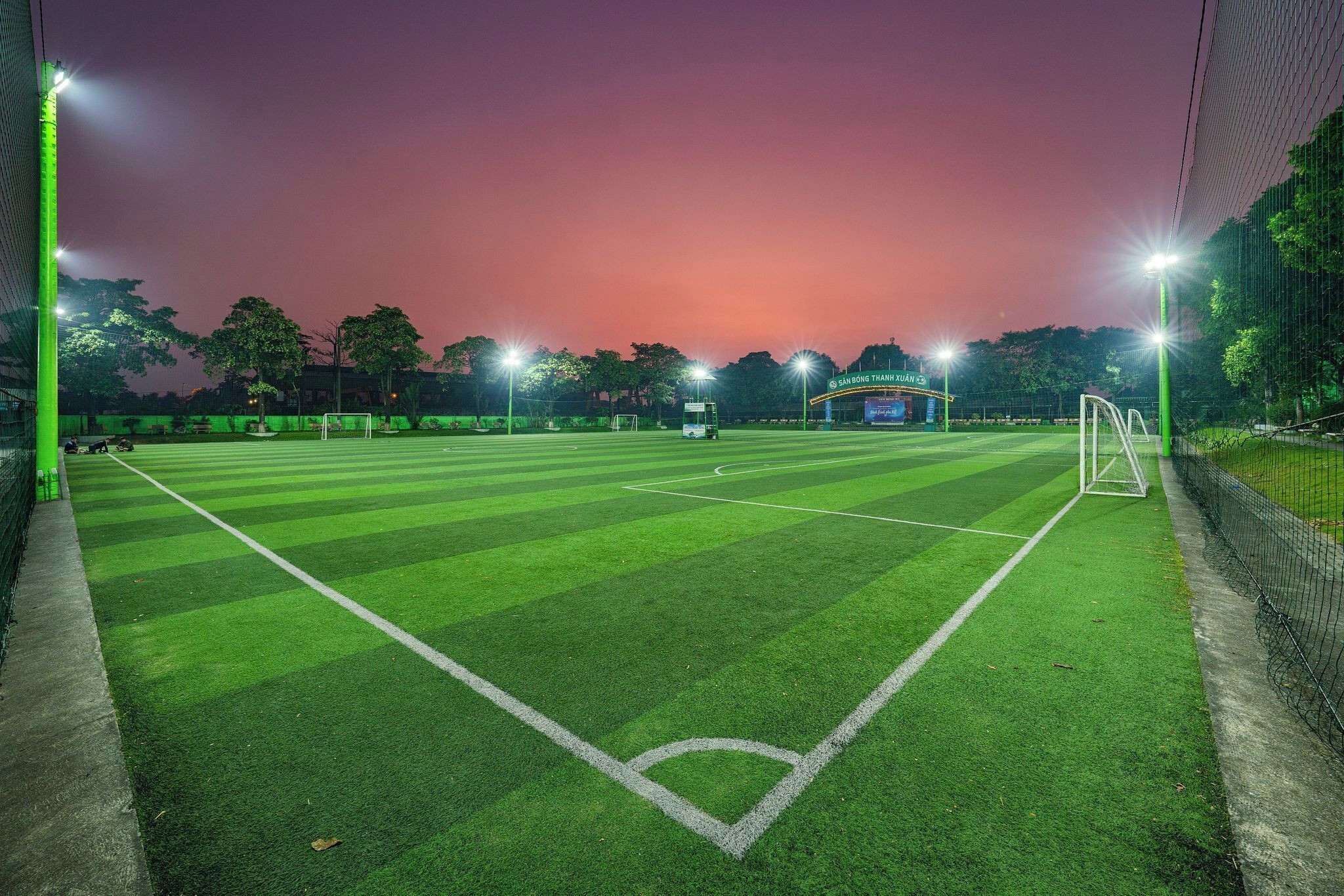 A well-lit soccer field at dusk with goalposts and a bus in the background.