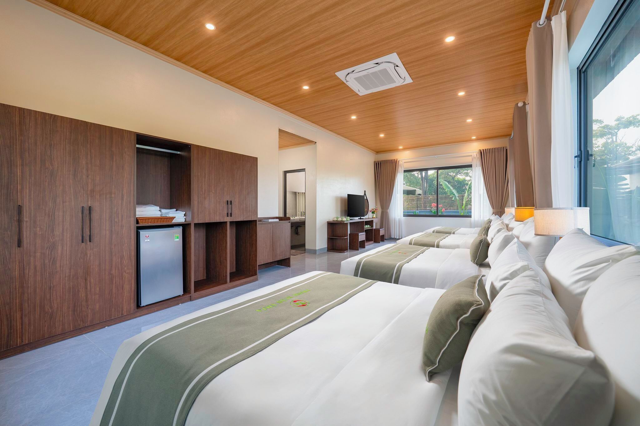 A modern hotel room with a wooden ceiling, a large window with curtains, a bed with a green bedspread, and a small kitchenette.