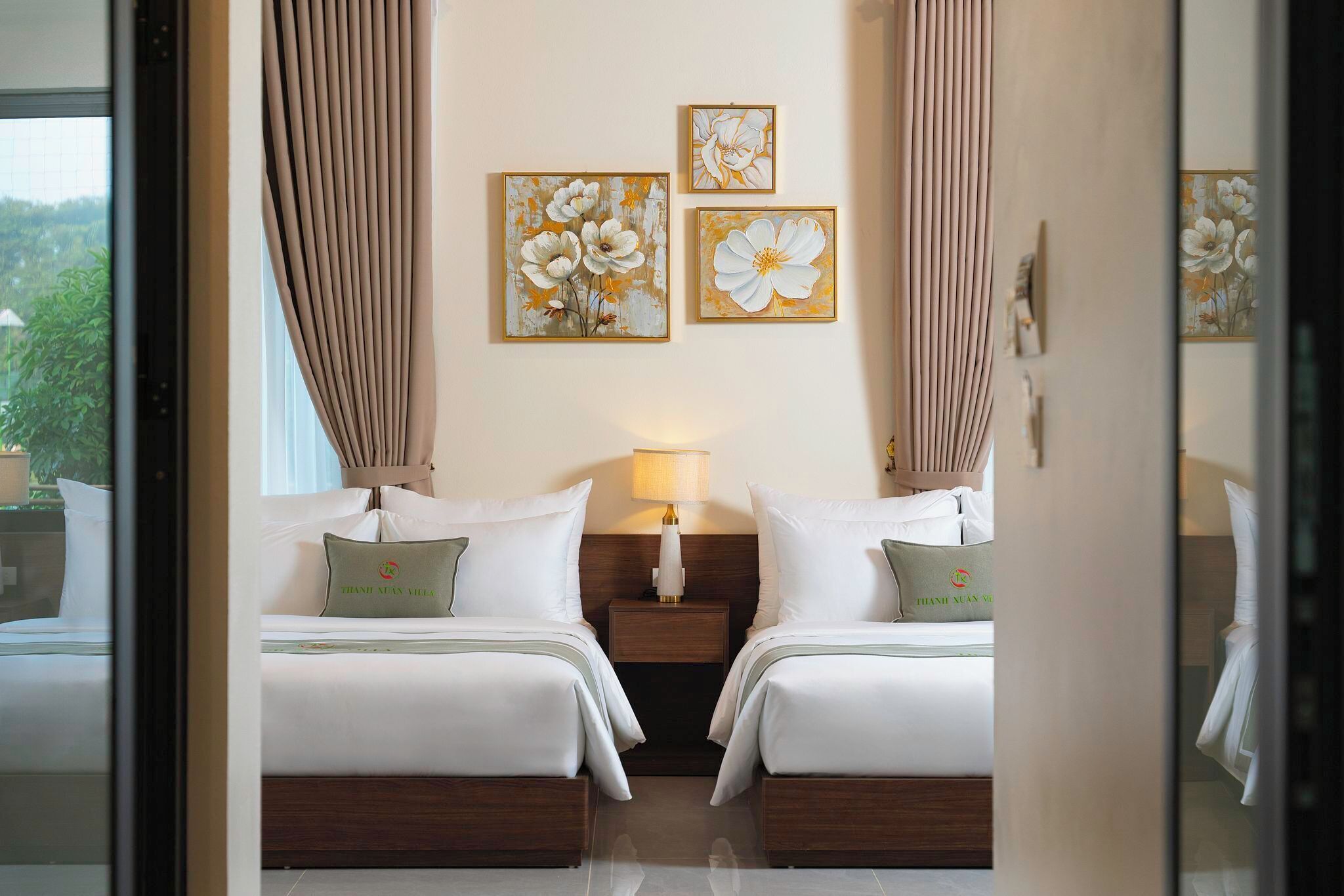 A hotel room with two beds, a nightstand with a lamp, and three floral paintings on the wall.