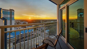 Ein Balkon mit Blick auf einen Sonnenuntergang über einer Stadtlandschaft, ein Stuhl und eine Glastür, die ein Gebäude reflektiert.