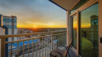 A balcony with a view of a sunset over a cityscape, a chair, and a glass door reflecting a building.