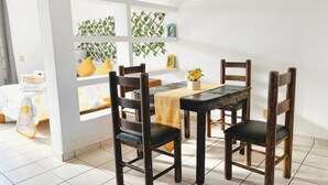 A dining area with a table and four chairs, a window with a plant, and a bed with a yellow pillow. - Mexican Cozy Apartment (Puerto Vallarta)